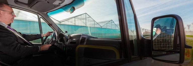 Man driving a van, his reflection in the side mirror captures a contemplative gaze. In the background, greenhouses stand tall, mirroring past journeys recorded like motor vehicle records.