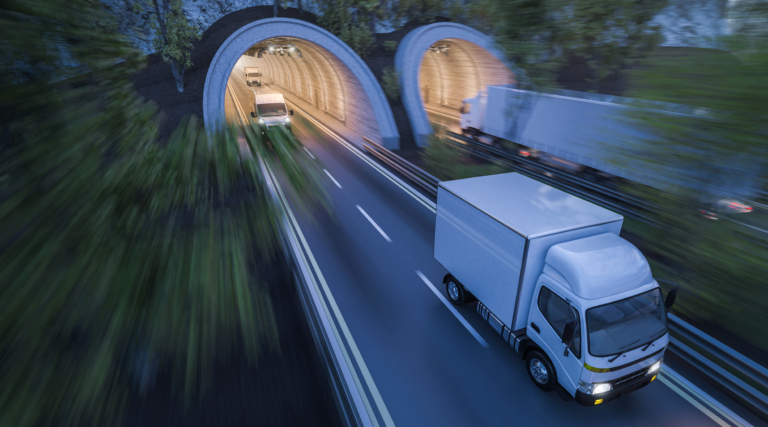 A white truck, likely monitored by Motor Vehicle Records, exits a tunnel on the highway. More trucks follow as greenery lines the road and another tunnel looms ahead.